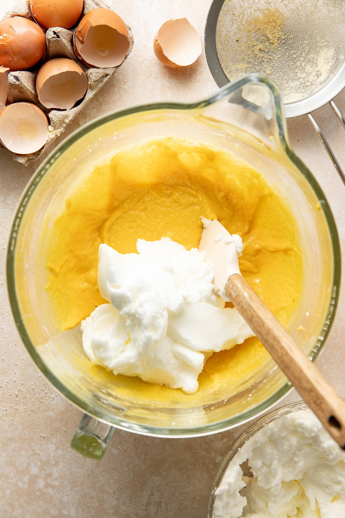 A glass mixing bowl with yellow batter and a dollop of whipped egg whites, a spatula, empty eggshells, and a metal strainer nearby on a countertop.