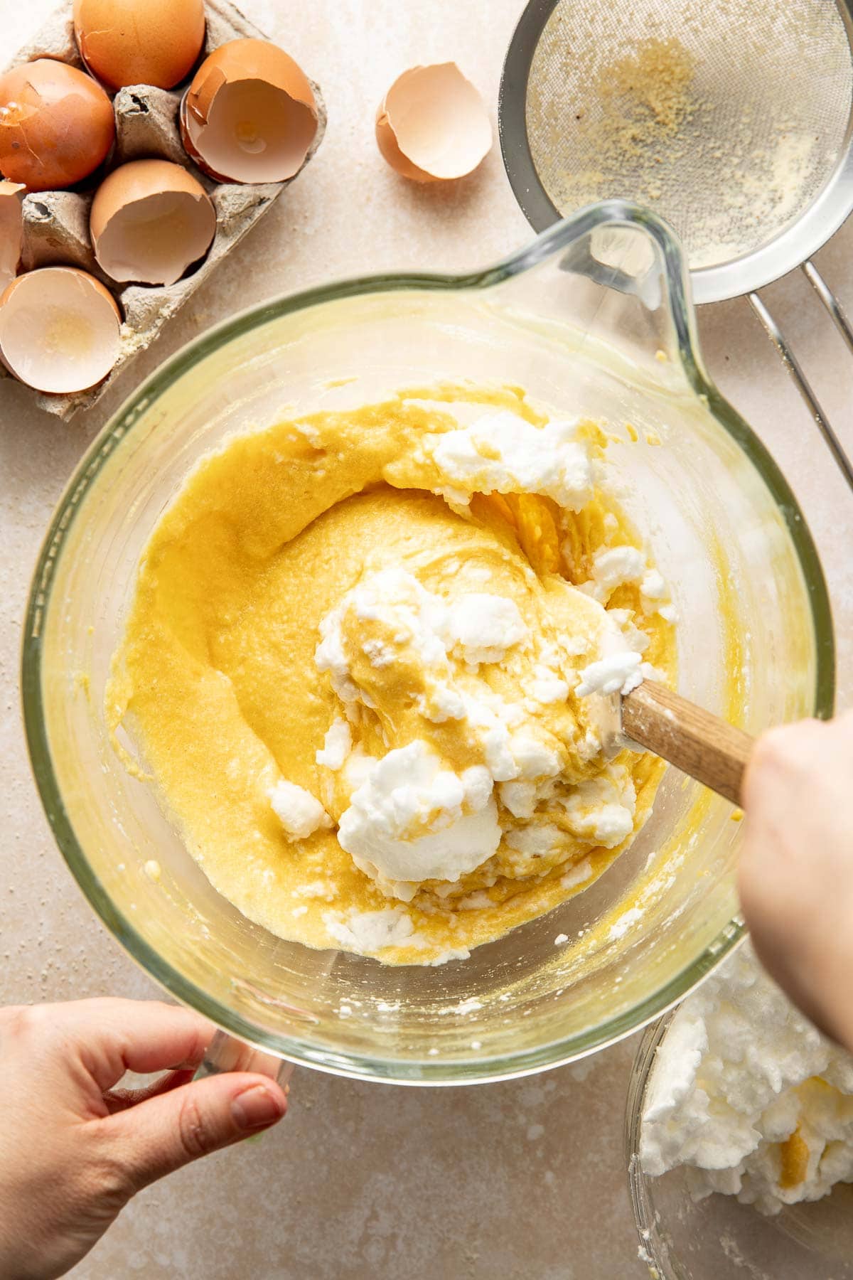 A hand mixes fluffy egg whites into a yellow batter in a glass bowl, with cracked eggshells, a sifter, and a carton of eggs nearby on a light countertop.