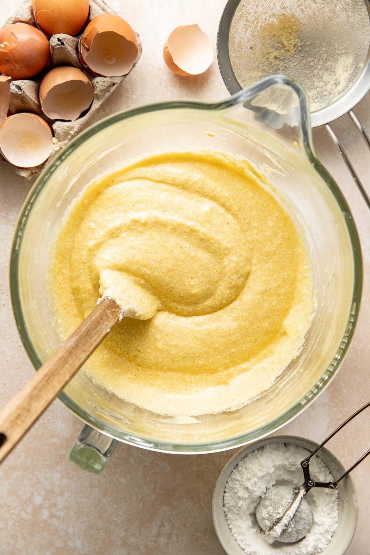 A glass mixing bowl with pale yellow cake batter being stirred by a wooden spoon. Around it are cracked eggshells in a carton and a small bowl of flour with a sifter on a light countertop.