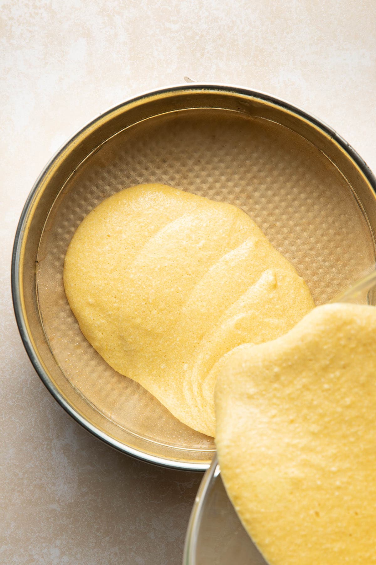A thick, yellow cake batter is being poured into a round, metal baking pan lined with parchment paper.