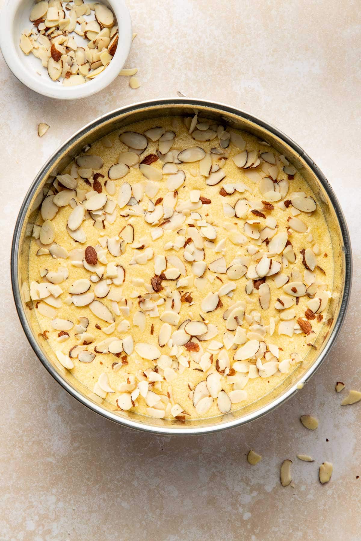 A round baking pan filled with batter, topped with sliced almonds, sits on a light-colored surface. A small bowl with more sliced almonds is nearby, with some almond slices scattered around.