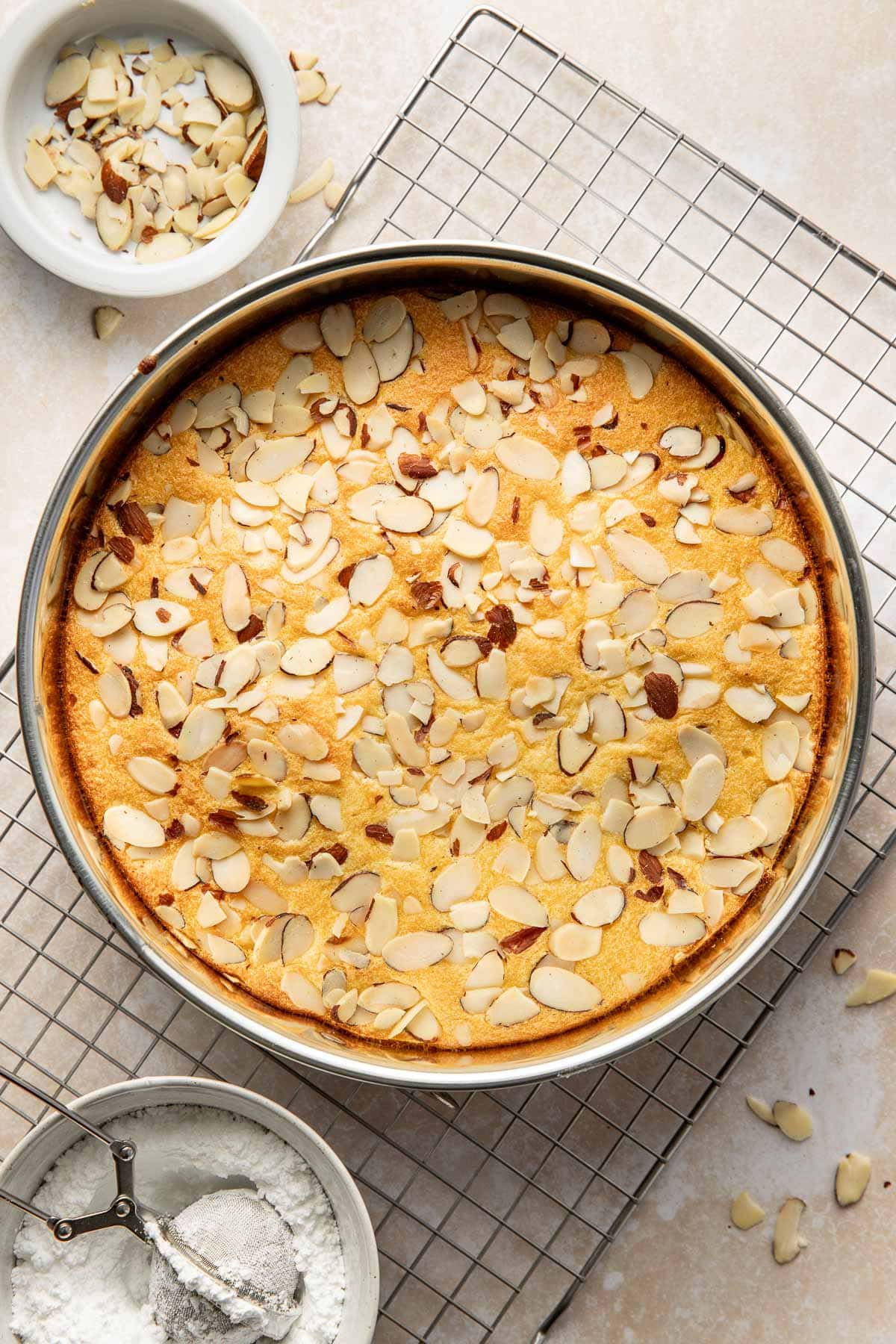 A round almond cake topped with sliced almonds in a baking pan cools on a wire rack, with bowls of sliced almonds and powdered sugar nearby.
