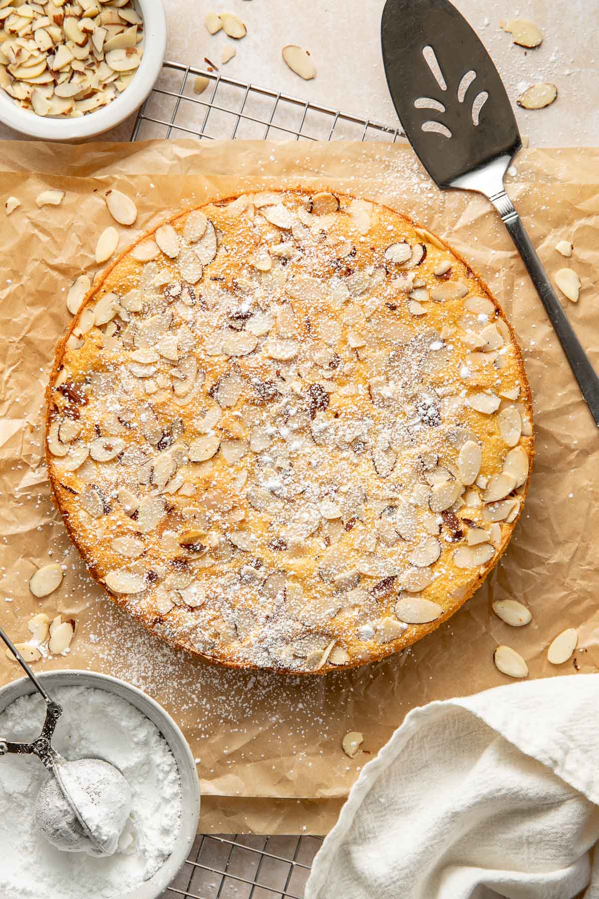 A round almond cake topped with sliced almonds and powdered sugar sits on parchment paper. Nearby are a pie server, a bowl of sliced almonds, a bowl of powdered sugar, and a white cloth napkin.