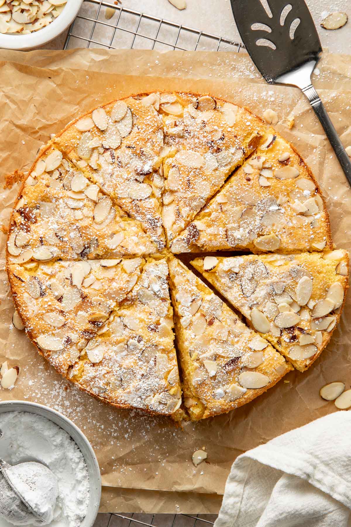 A round almond cake topped with sliced almonds and powdered sugar, sliced into eight pieces, sits on parchment paper with a pie server, a small bowl of powdered sugar, and a white cloth nearby.
