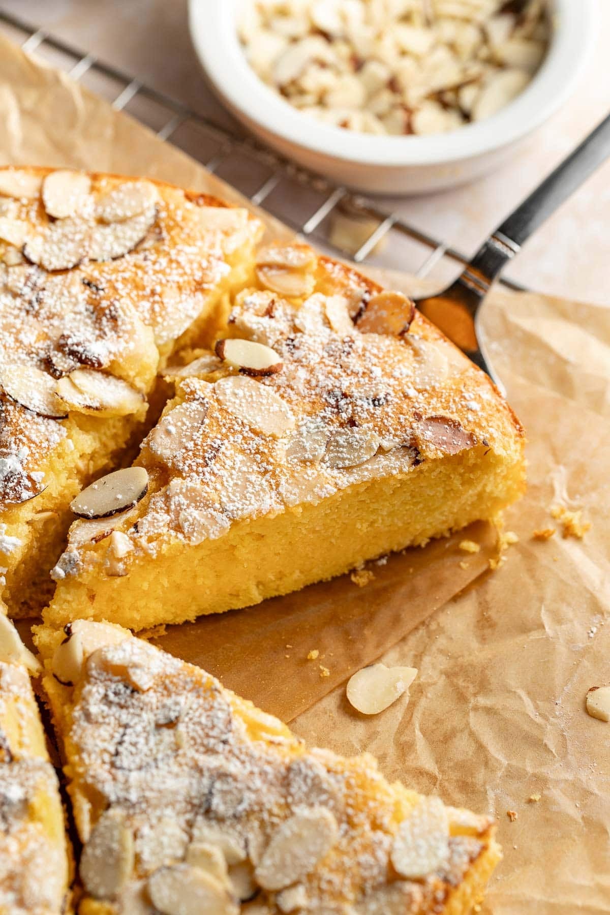 A close-up of a sliced almond cake on parchment paper, topped with powdered sugar and almond flakes. A small bowl with more almond slices is visible in the background.