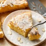 A slice of almond cake topped with powdered sugar and almond flakes sits on a speckled plate, with a serving utensil lifting a piece. The background shows more cake and scattered almond slices.