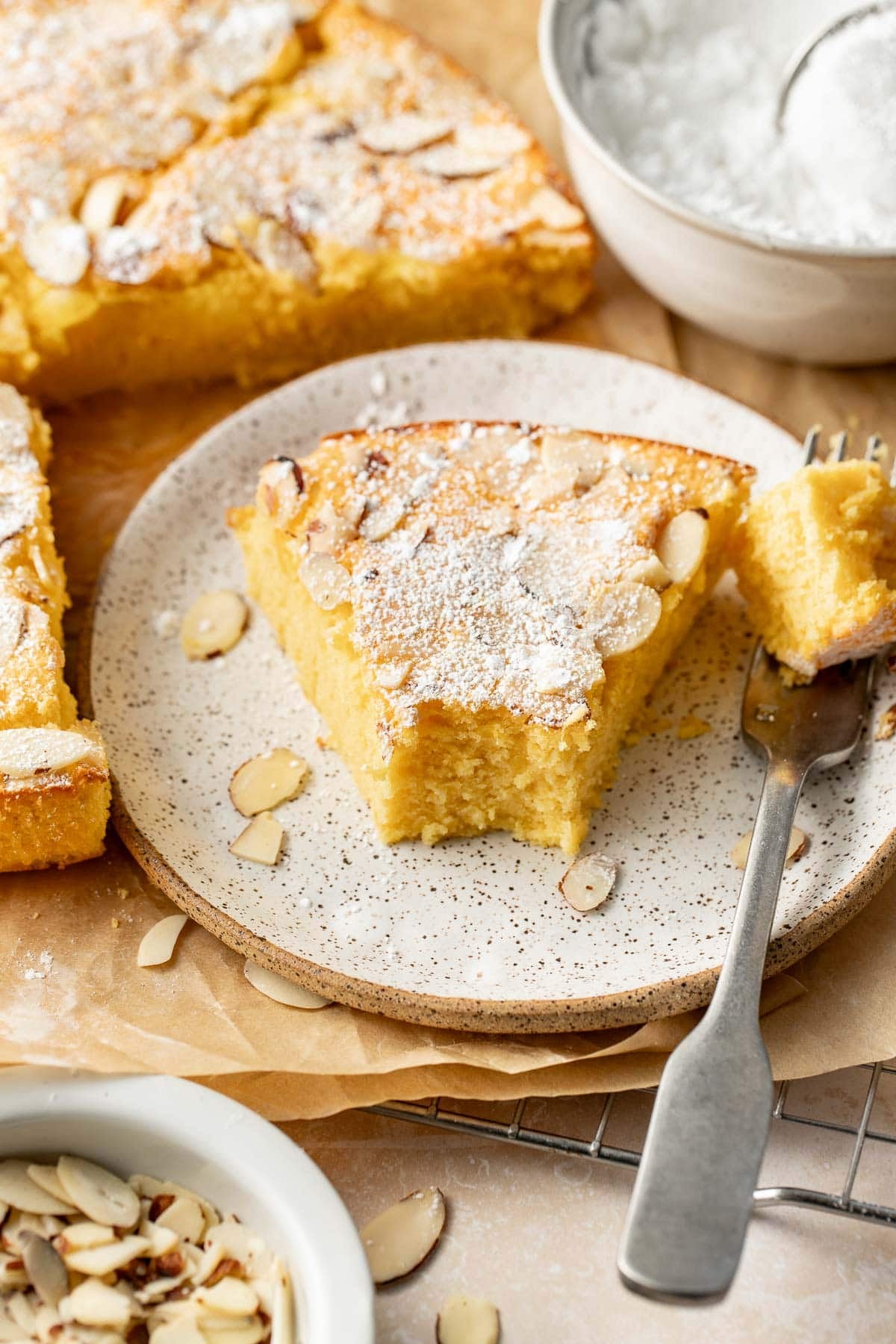 A slice of almond cake topped with powdered sugar and almond flakes sits on a plate, with a fork holding a bite-sized piece. More cake, a bowl of powdered sugar, and almond slices are nearby.