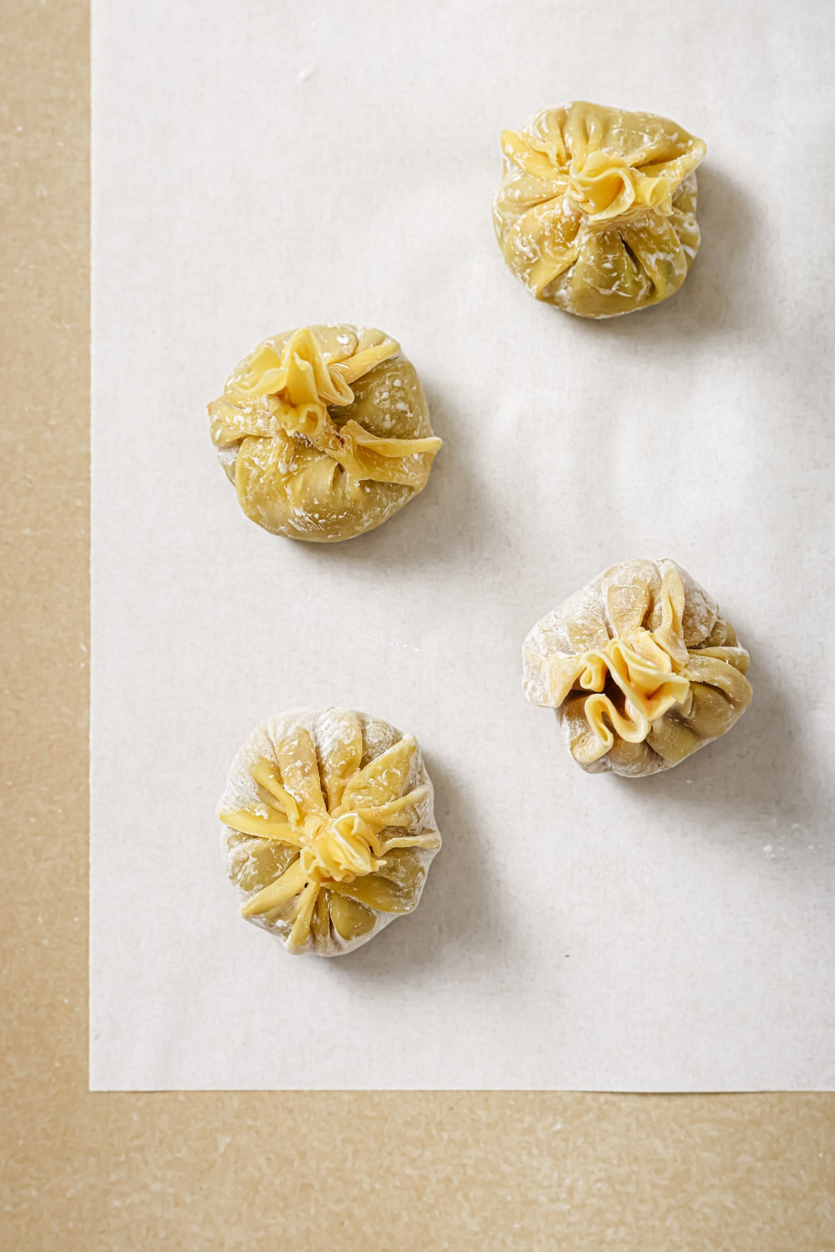 Four uncooked dumplings with pleated tops sit on a sheet of white parchment paper, placed on a beige surface. The dumplings are dusted lightly with flour and arranged in a loose square pattern.