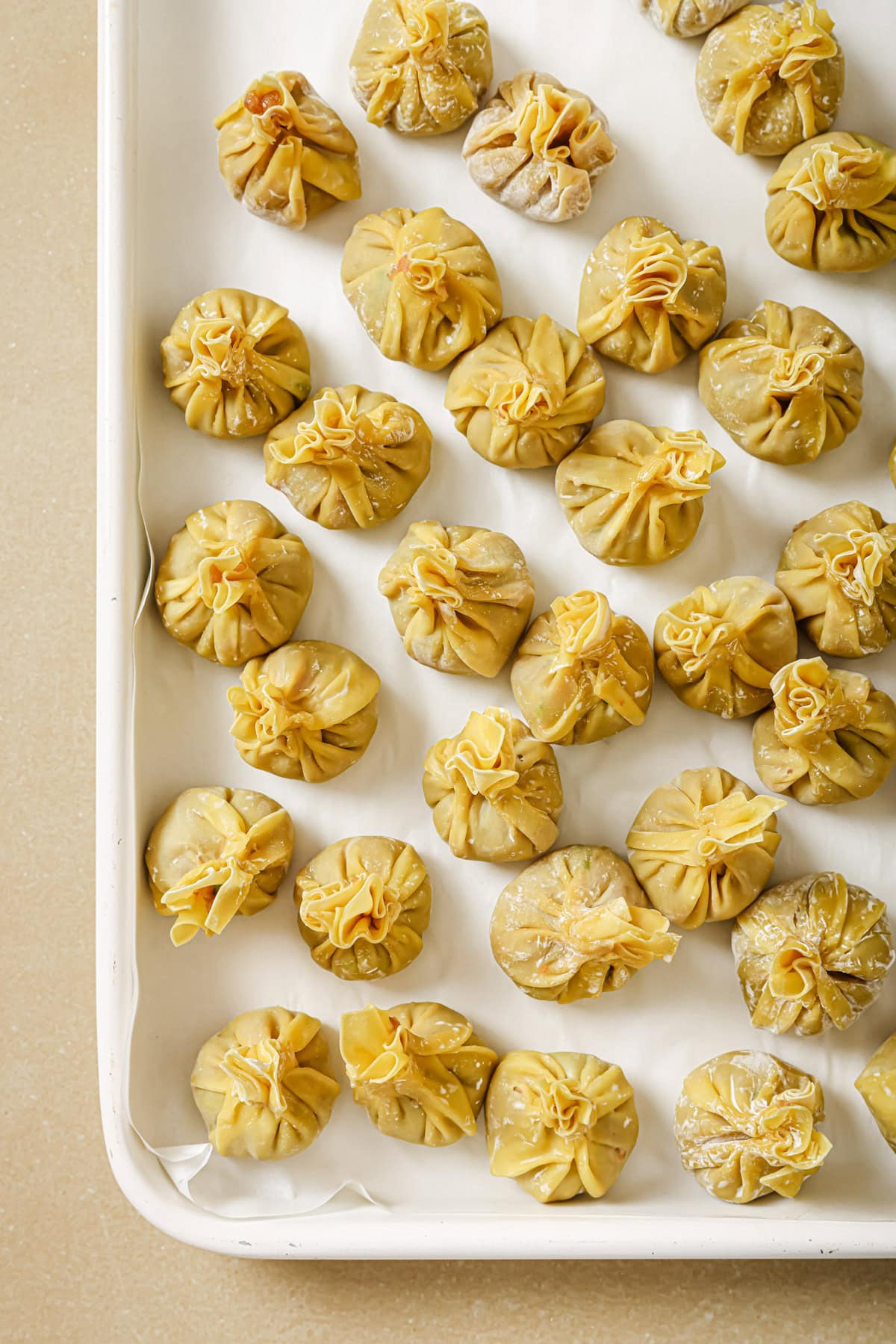 A baking tray lined with parchment paper, filled with neatly arranged, uncooked dumplings shaped into small, round pouches with gathered tops.