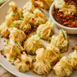 A plate of steamed dumplings garnished with sesame seeds and sliced green onions, served with a bowl of soy-based dipping sauce with chili flakes.