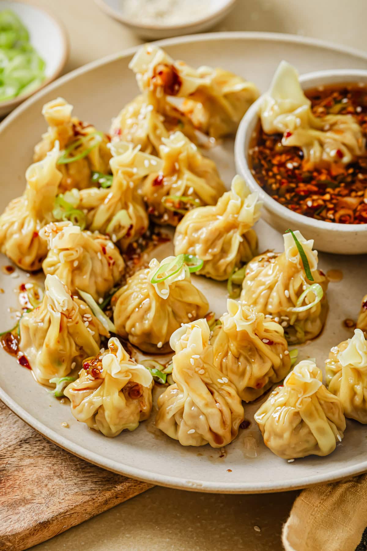 A plate of steamed dumplings garnished with sesame seeds and sliced green onions, served with a bowl of soy-based dipping sauce with chili flakes.