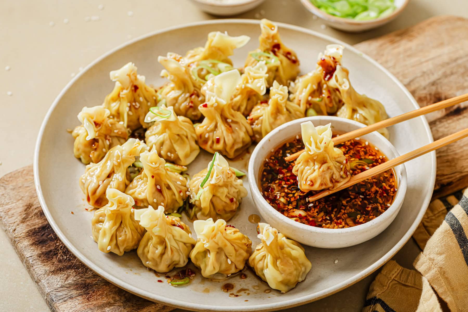 A plate of steamed dumplings arranged around a bowl of spicy dipping sauce, with one dumpling being held by chopsticks over the sauce; some chopped green onions and sesame seeds are sprinkled on top.