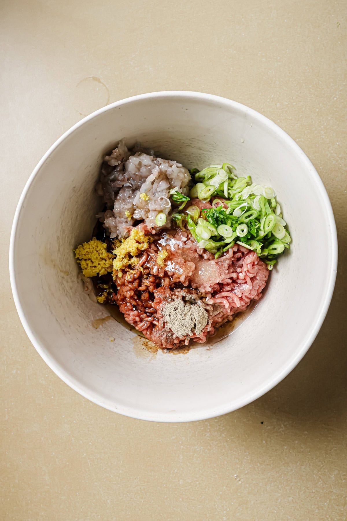 A white mixing bowl containing raw ground meat, chopped shrimp, sliced green onions, grated ginger, minced garlic, soy sauce, and spices on a beige surface.