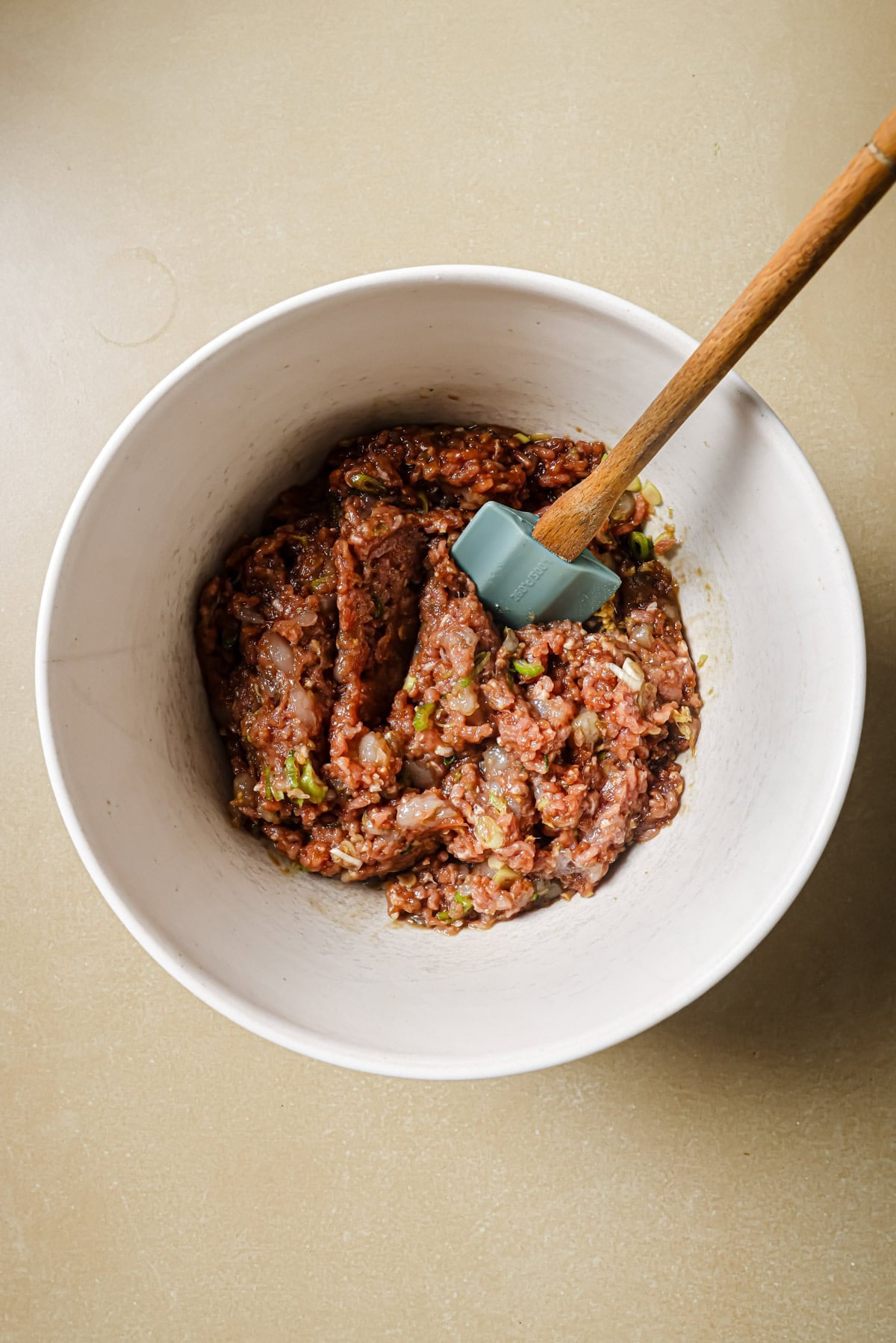 A white mixing bowl filled with seasoned raw ground meat and chopped vegetables, being stirred with a blue spatula that has a wooden handle, set on a beige countertop.