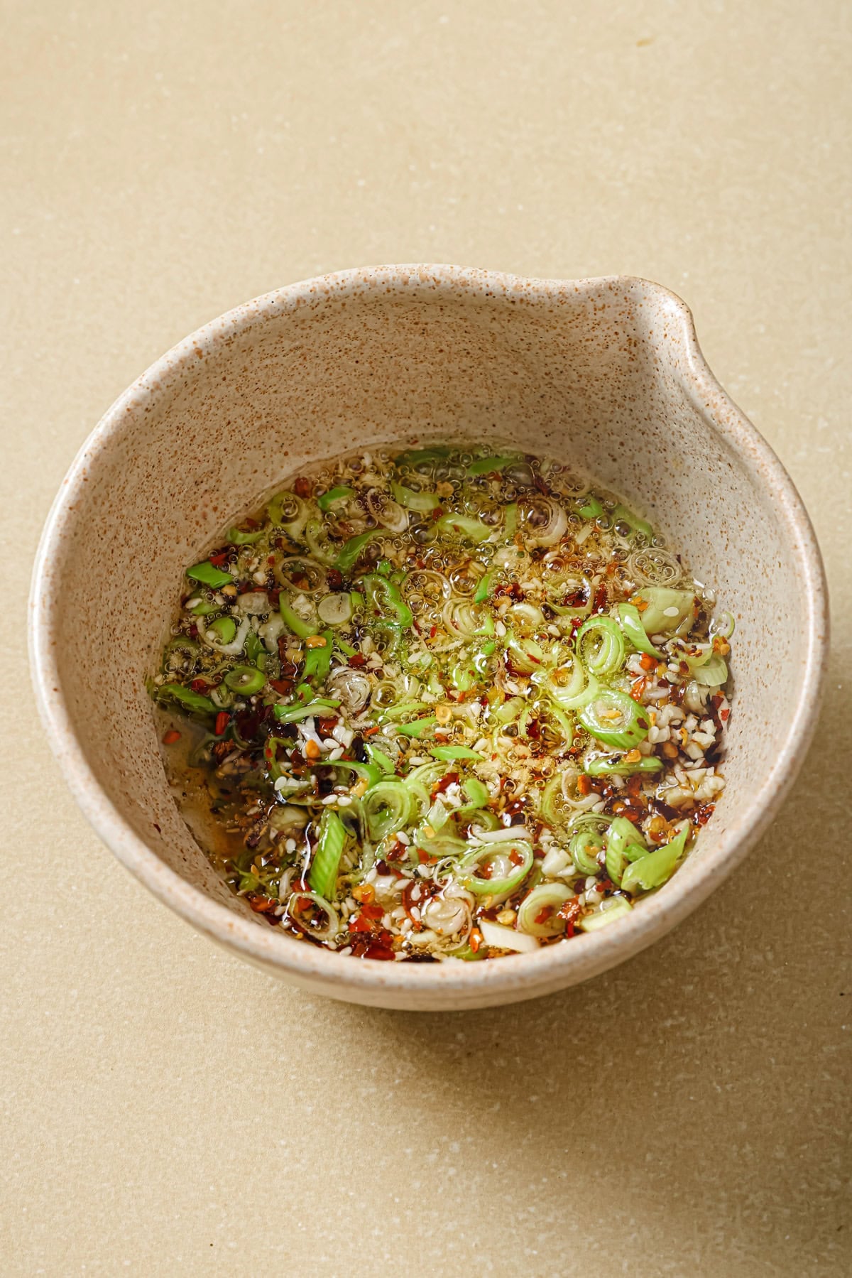 A speckled ceramic bowl filled with a mixture of sliced green onions, chili flakes, minced garlic, and oil on a beige countertop.