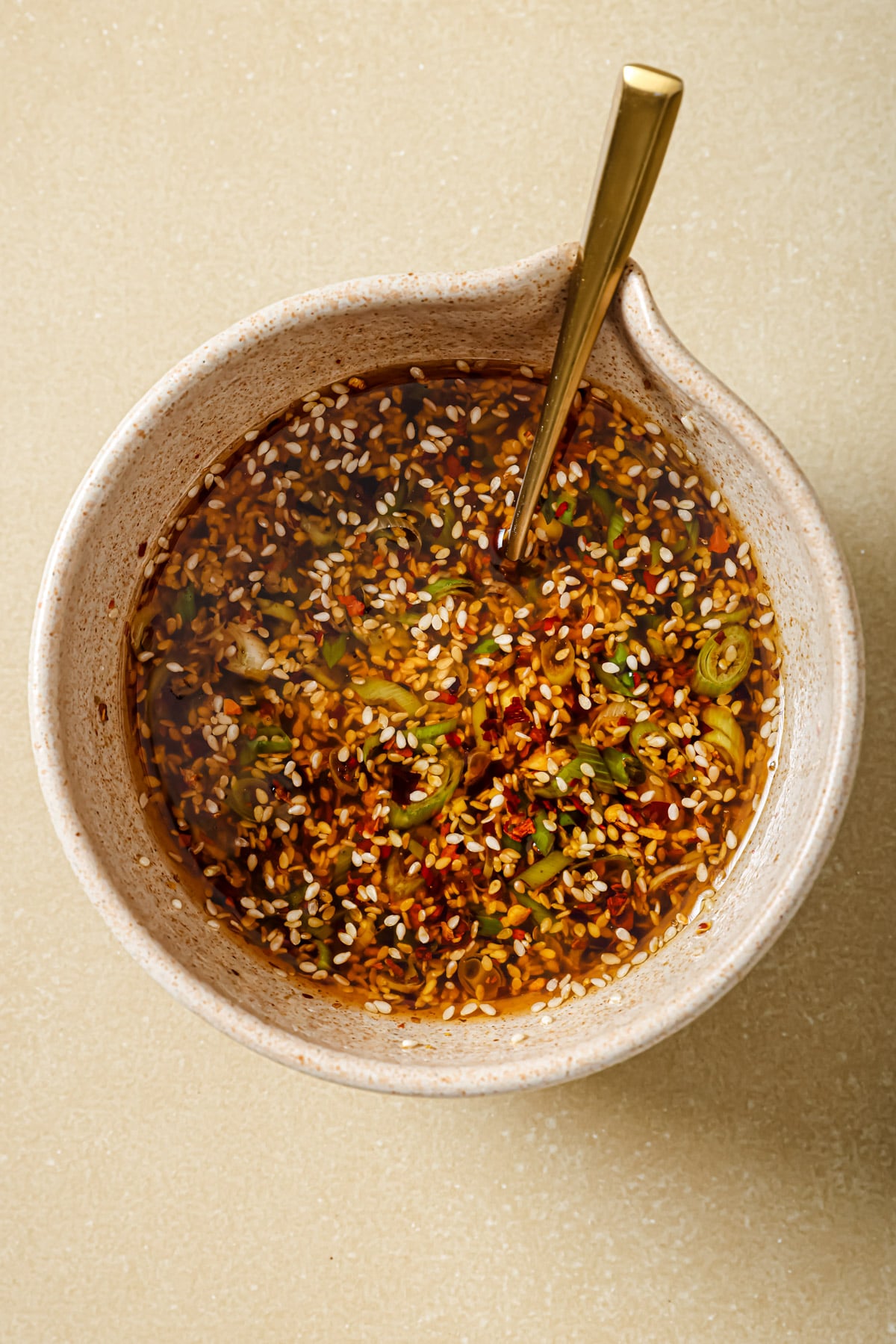 A beige bowl filled with a colorful sauce containing sliced green onions, sesame seeds, and chili flakes, with a gold spoon resting inside. The bowl sits on a light tan surface.