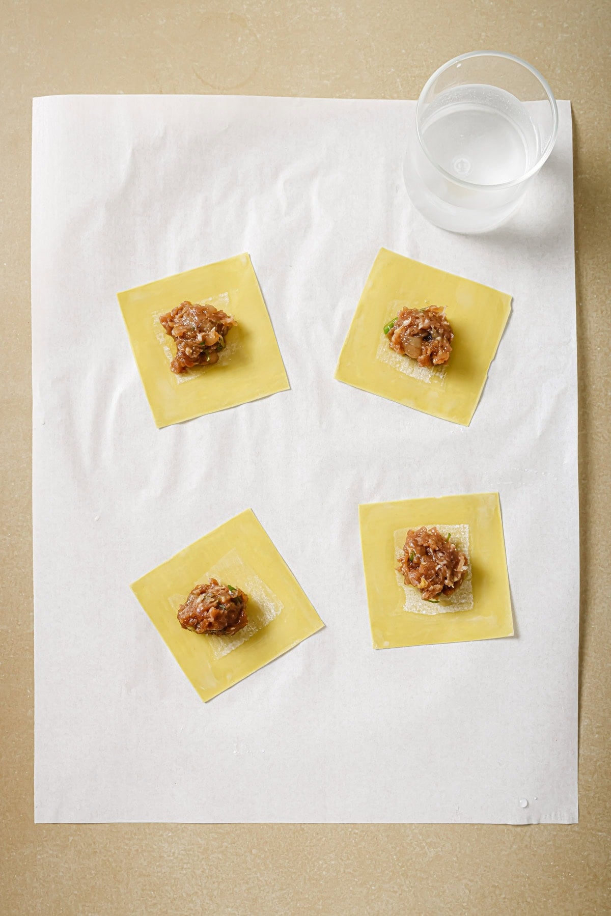 Four yellow wonton wrappers each topped with a small mound of meat filling are arranged on a sheet of parchment paper; a clear plastic cup of water sits in the top right corner.