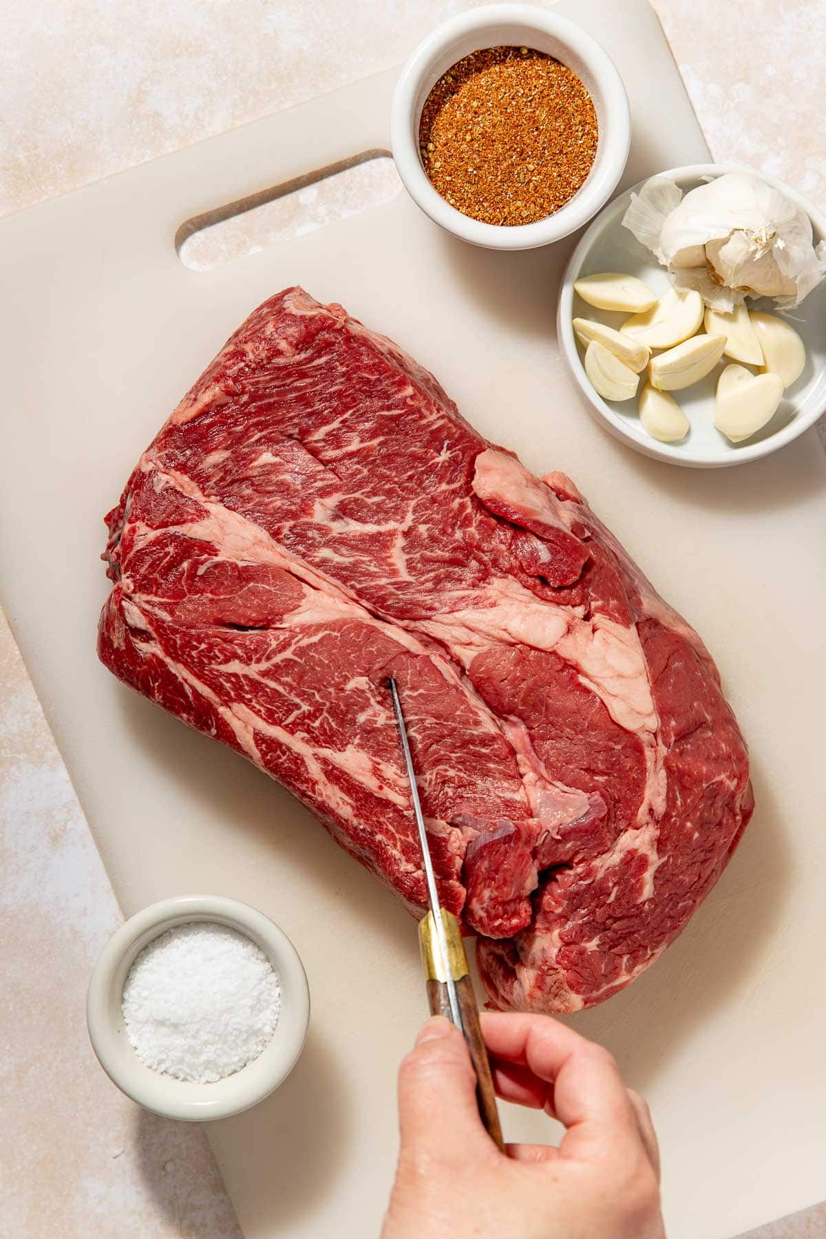 A hand holds a knife, slicing into a large raw beef roast on a cutting board. Nearby are small bowls containing salt, spice rub, and peeled garlic cloves.