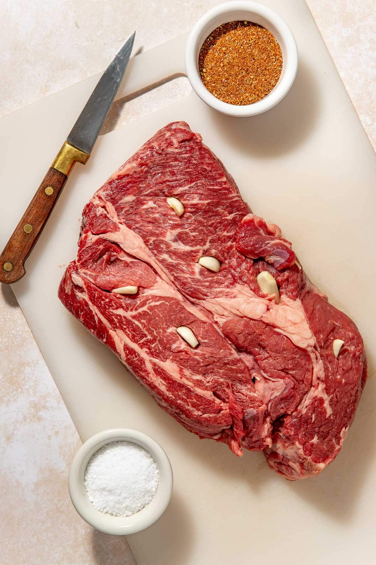 A raw beef brisket with garlic cloves inserted sits on a cutting board next to a knife, a small bowl of seasoning, and a bowl of salt.