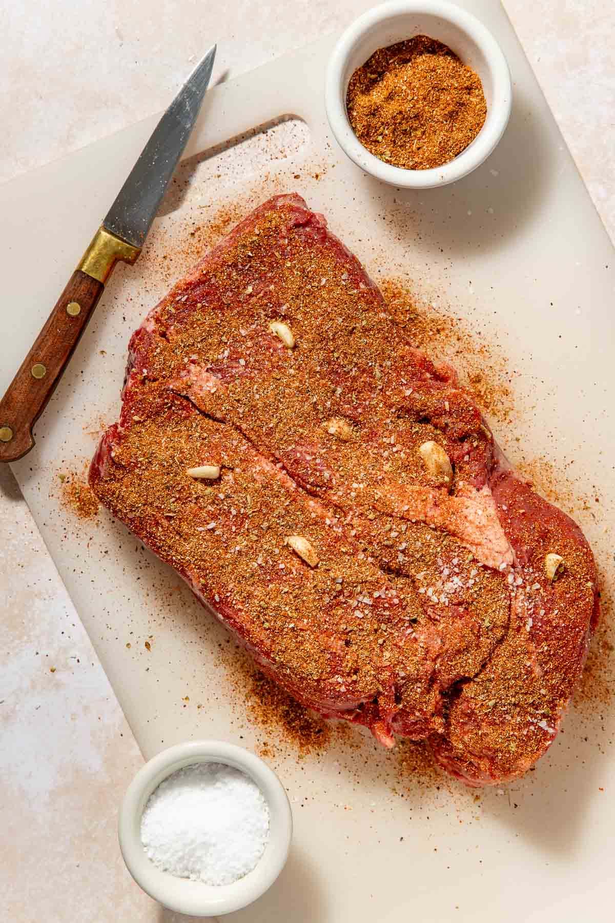 A raw seasoned beef roast with garlic cloves is on a cutting board next to a knife, a small bowl of spice rub, and a bowl of salt. The roast is coated with a dry spice mix.