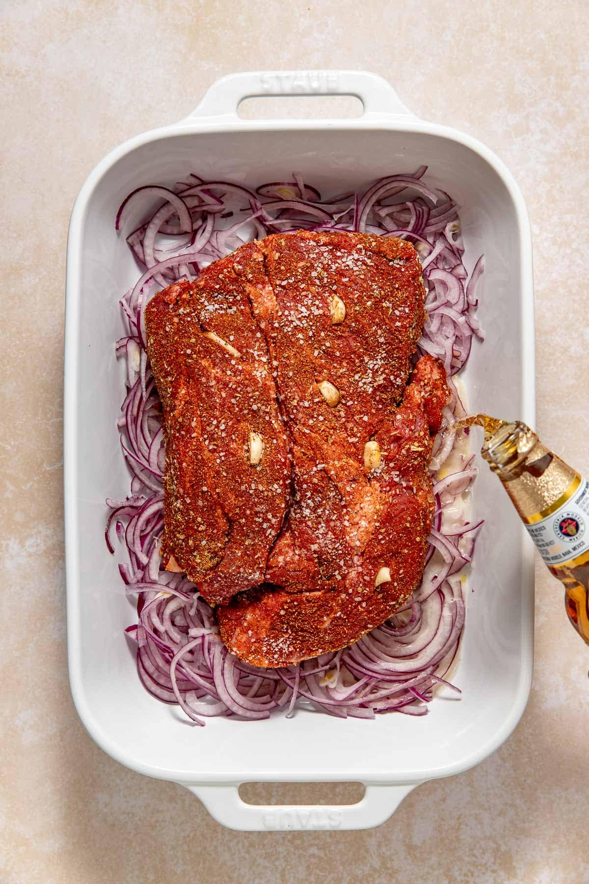 A white baking dish with sliced red onions, a seasoned raw beef roast topped with garlic cloves, and someone pouring beer from a bottle over the meat.