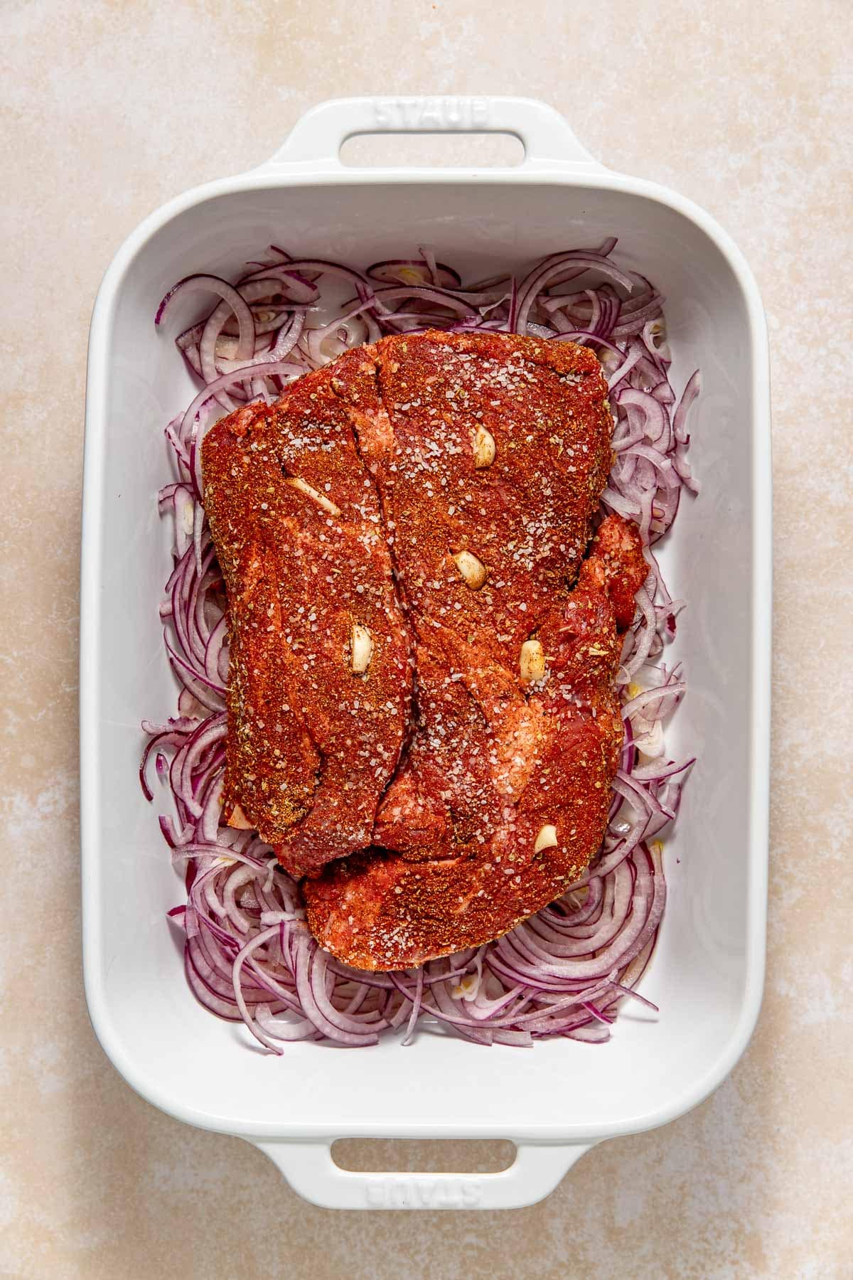 A white baking dish filled with sliced red onions topped with a large piece of seasoned raw meat with visible garlic pieces.