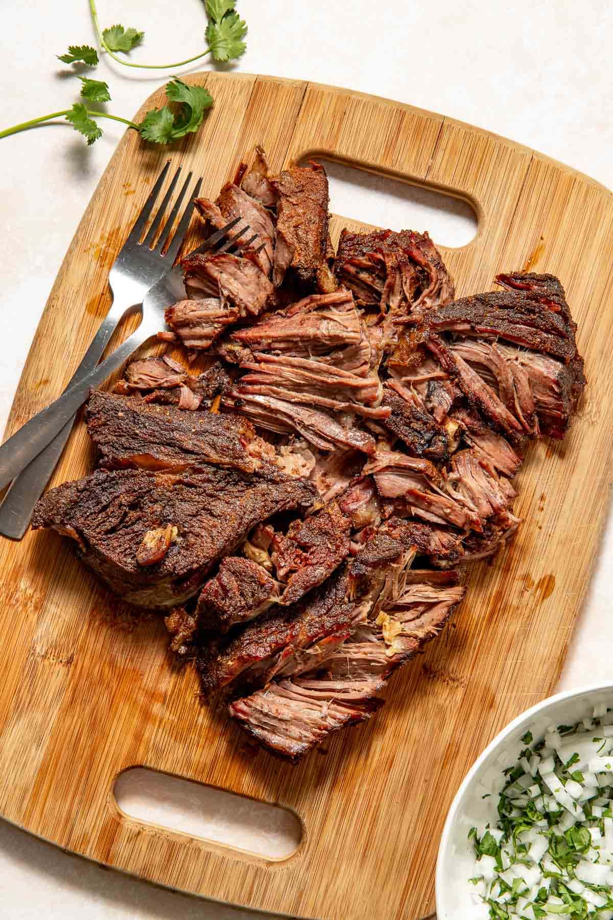 Shredded, seasoned beef is arranged on a wooden cutting board next to a fork and knife, with cilantro and a small bowl of chopped onions and herbs nearby.