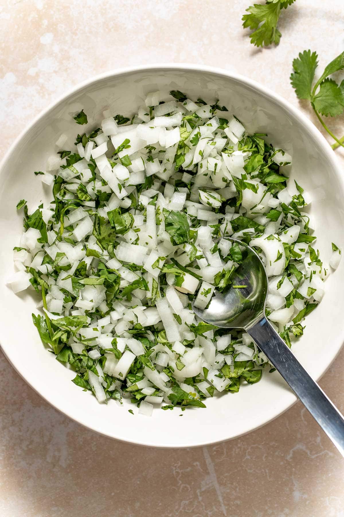 A white bowl filled with chopped white onions and fresh cilantro, with a metal spoon inside. A sprig of cilantro is on the beige surface beside the bowl.