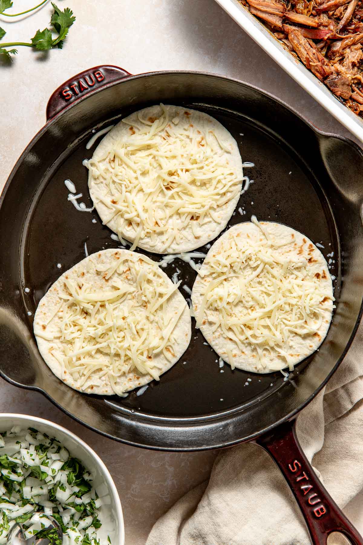 Three tortillas topped with shredded cheese are warming in a cast iron skillet. Surrounding the skillet are bowls of chopped onions, cilantro, and a dish of shredded meat.