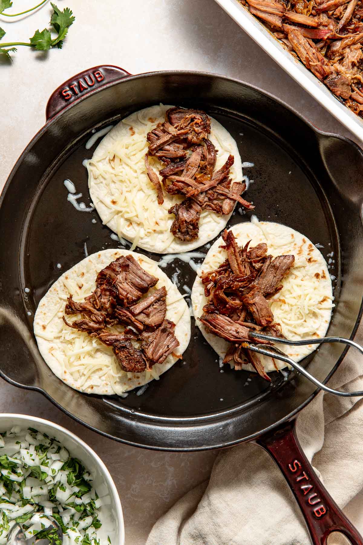 A cast iron skillet with three tortillas topped with shredded cheese and braised beef. Tongs are arranging the beef. Surrounding the skillet are a dish of chopped onions and cilantro, and a tray of more shredded beef.