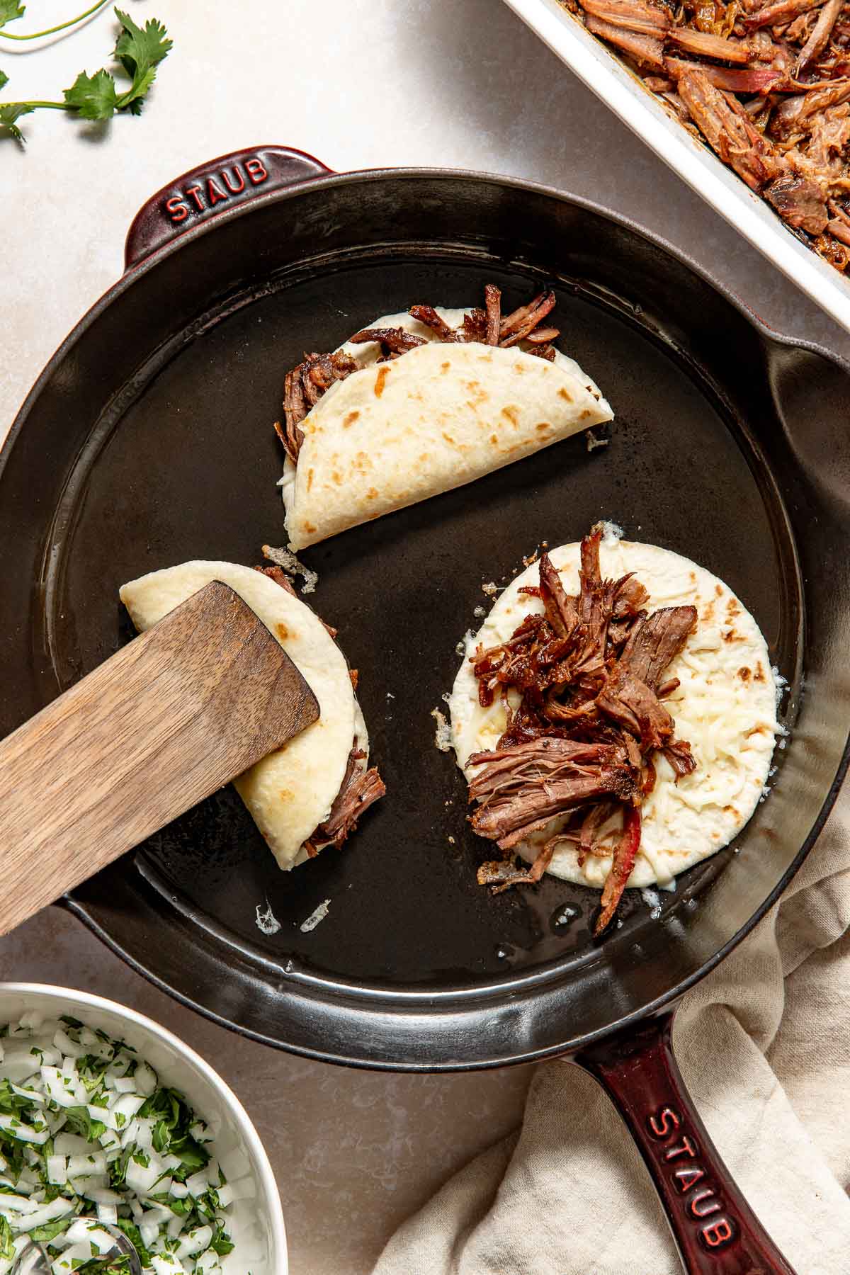 A cast iron skillet with two quesadillas being cooked; each stuffed with shredded beef and melted cheese. A spatula lifts one quesadilla. Bowls of chopped onion, cilantro, and extra beef are nearby.