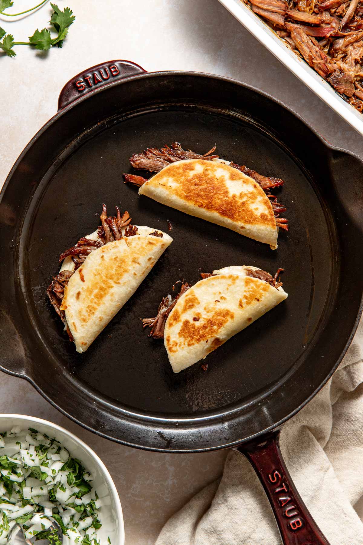 Two folded, golden-brown tacos filled with shredded meat cooking in a large black skillet. Fresh cilantro and chopped onions are in a white bowl nearby, with part of a casserole dish visible in the corner.
