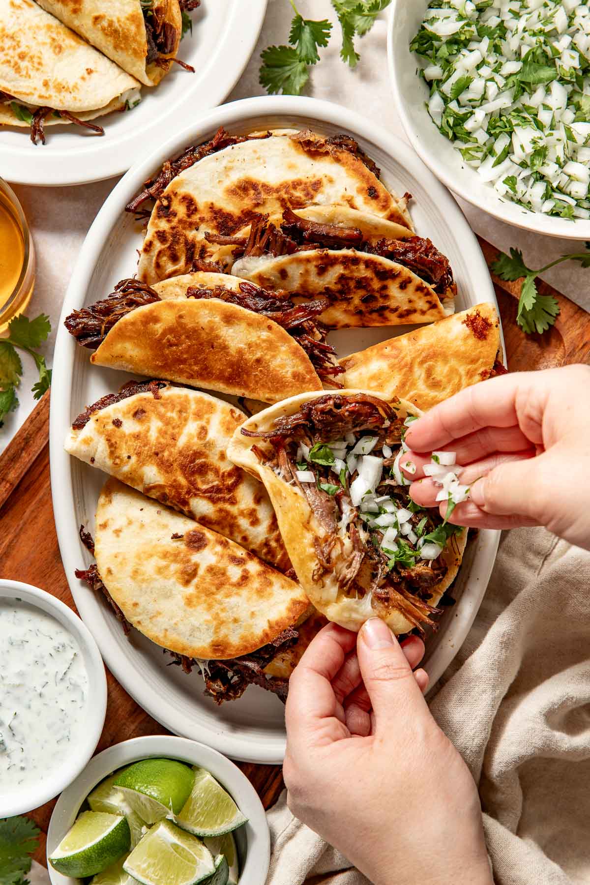 A person adds chopped onions and cilantro to a birria taco on a white plate, surrounded by more tacos, lime wedges, and bowls of chopped onions with cilantro and sauce.