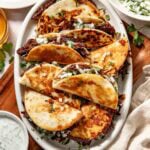 A white platter holds several crispy birria tacos filled with shredded meat, garnished with chopped onions and cilantro. Bowls of lime wedges, chopped onions, and dipping sauce surround the platter.