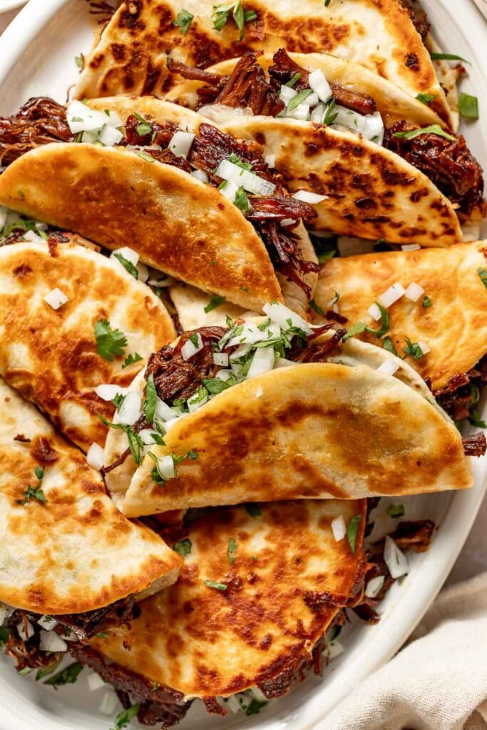 Crispy golden tortillas filled with shredded beef, topped with chopped onions and fresh cilantro, arranged on a white platter.
