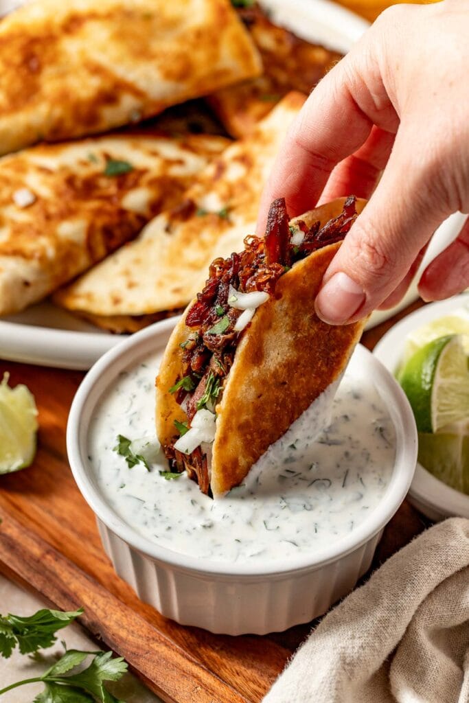 A hand dips a crispy, folded taco filled with shredded meat and chopped onions into a small bowl of creamy cilantro sauce, with more tacos and lime wedges in the background.