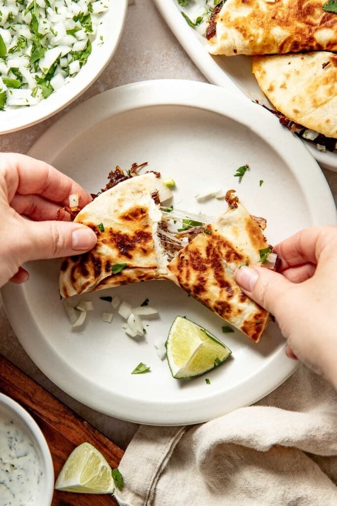 A person pulls apart a crispy taco filled with shredded meat on a white plate. Chopped onions, cilantro, and lime wedges are on the plate, with more toppings and tacos visible in the background.