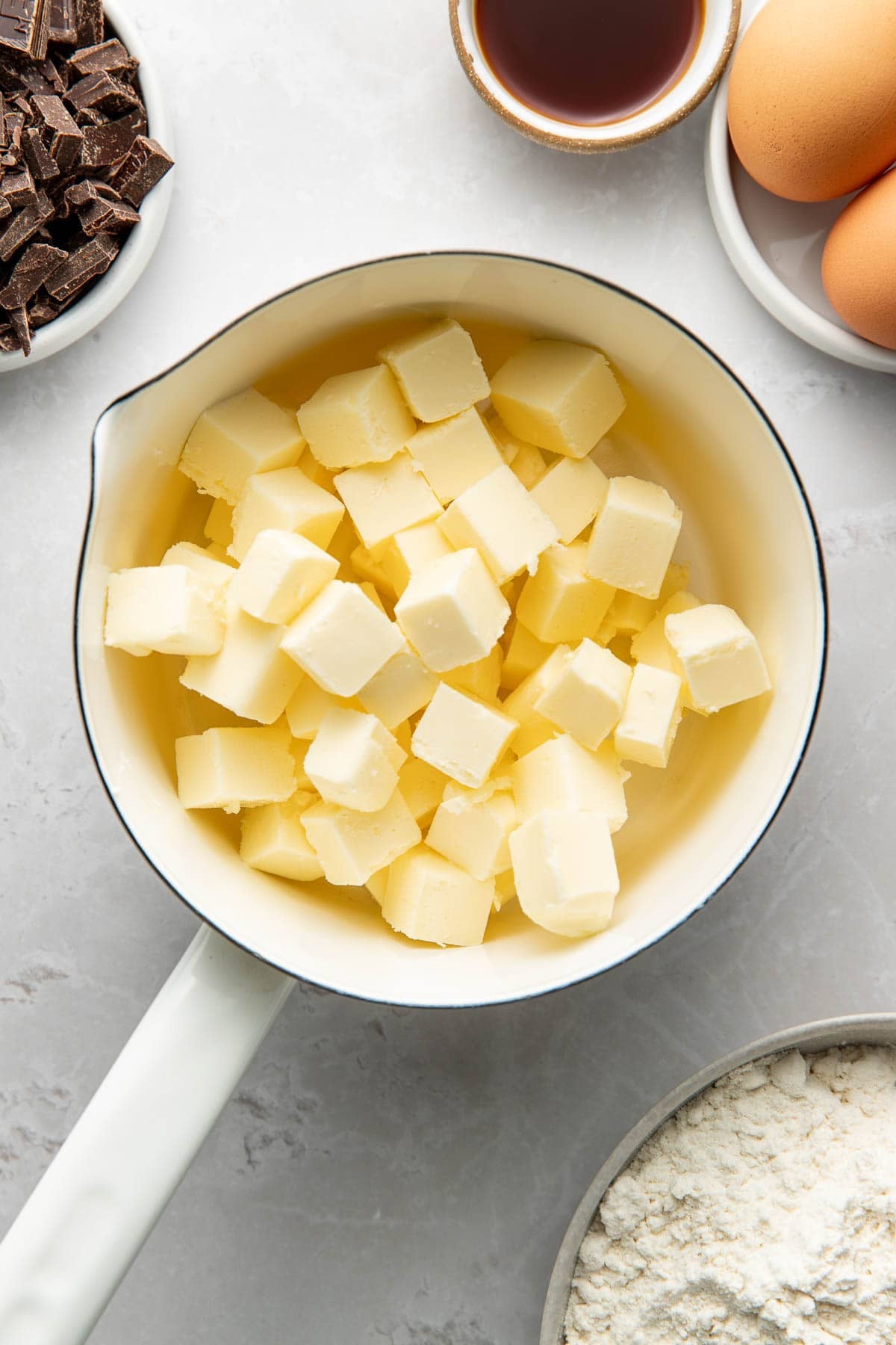 A white saucepan filled with cubed butter sits on a light countertop, surrounded by bowls containing chocolate chunks, vanilla extract, eggs, and flour.
