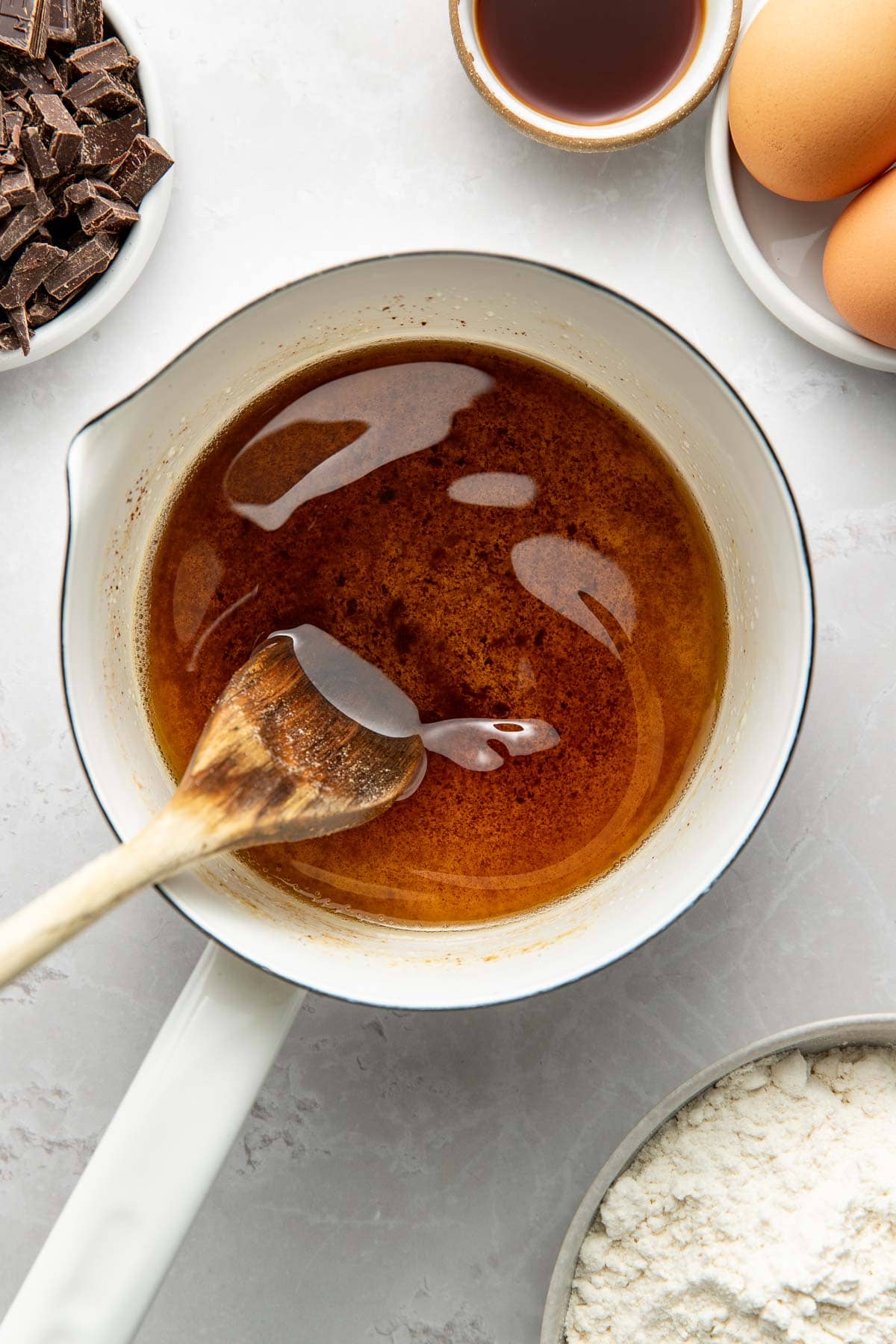 A saucepan with melted brown butter and a wooden spoon, surrounded by eggs, a bowl of flour, chopped chocolate, and a small bowl of vanilla extract on a light countertop.