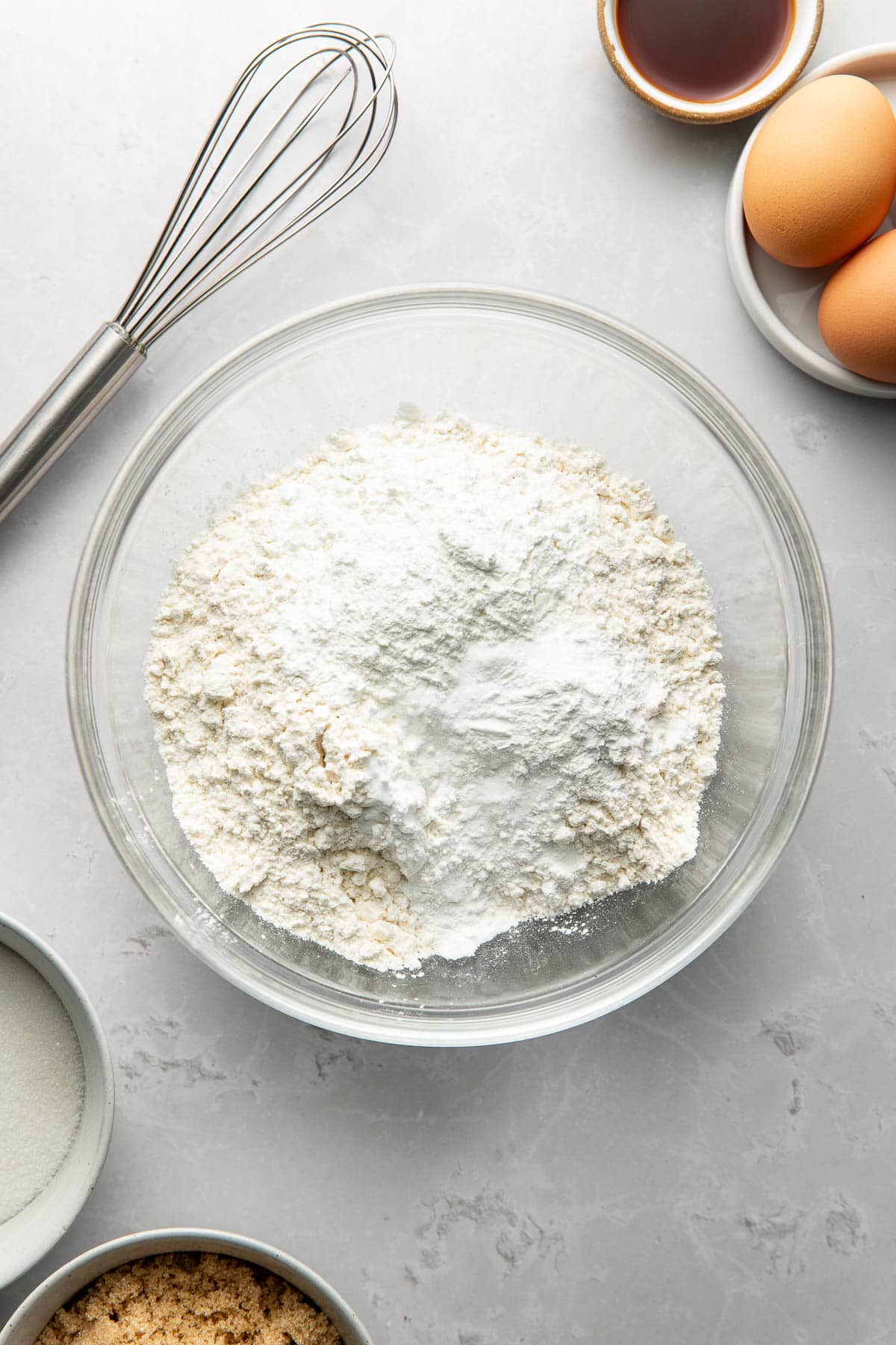 A glass bowl filled with flour sits on a countertop, surrounded by a whisk, two eggs, a small bowl of vanilla extract, and bowls of white and brown sugar.