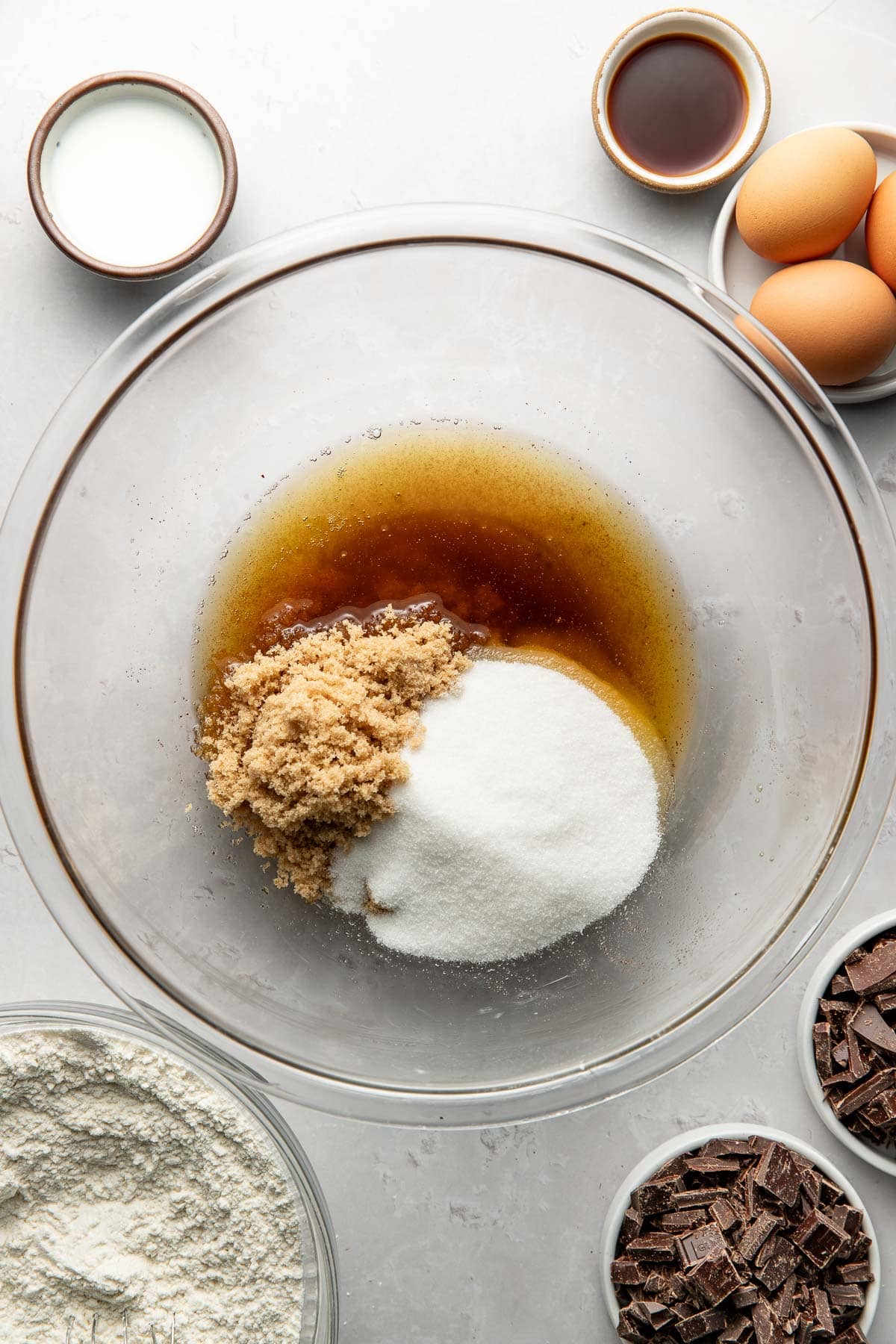 A glass bowl with brown sugar, white sugar, and melted butter sits on a counter, surrounded by bowls of flour, chocolate chunks, eggs, vanilla extract, and milk, all ready for baking.