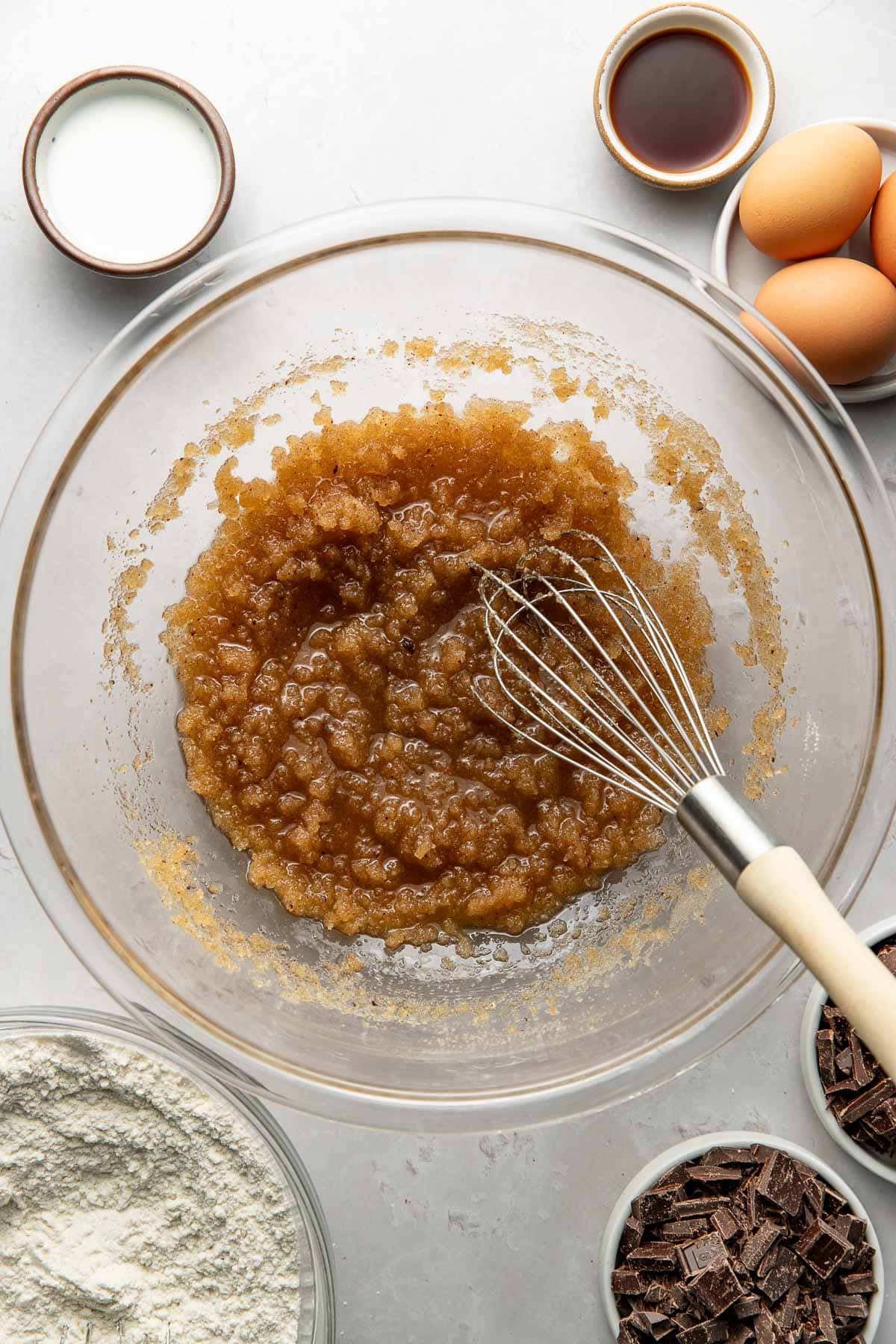 A glass mixing bowl with a whisk contains a brown sugar and melted butter mixture. Surrounding the bowl are small bowls with flour, chocolate chunks, milk, vanilla, and three eggs on a light surface.