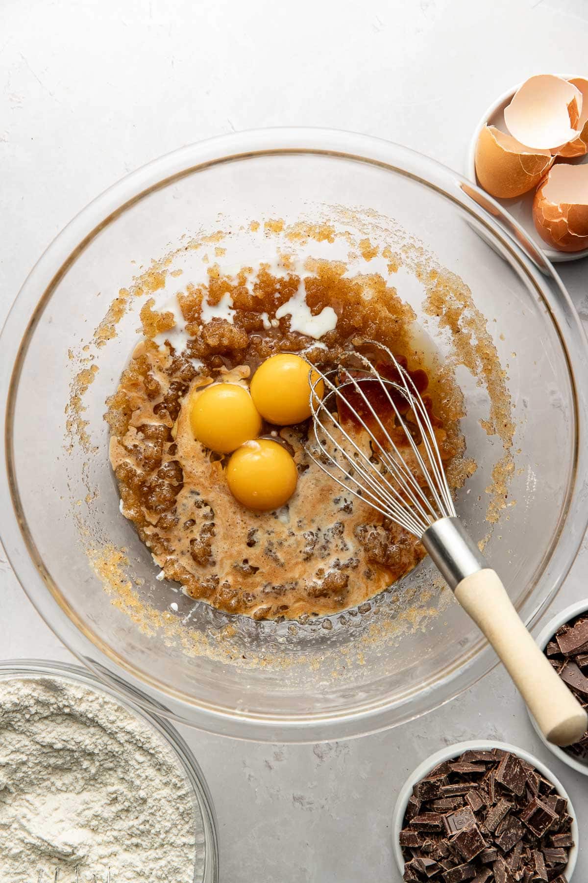 A glass bowl with three eggs, brown sugar, vanilla, and wet ingredients being whisked. Surrounding the bowl are containers with flour, chocolate chunks, and eggshells on a light surface.