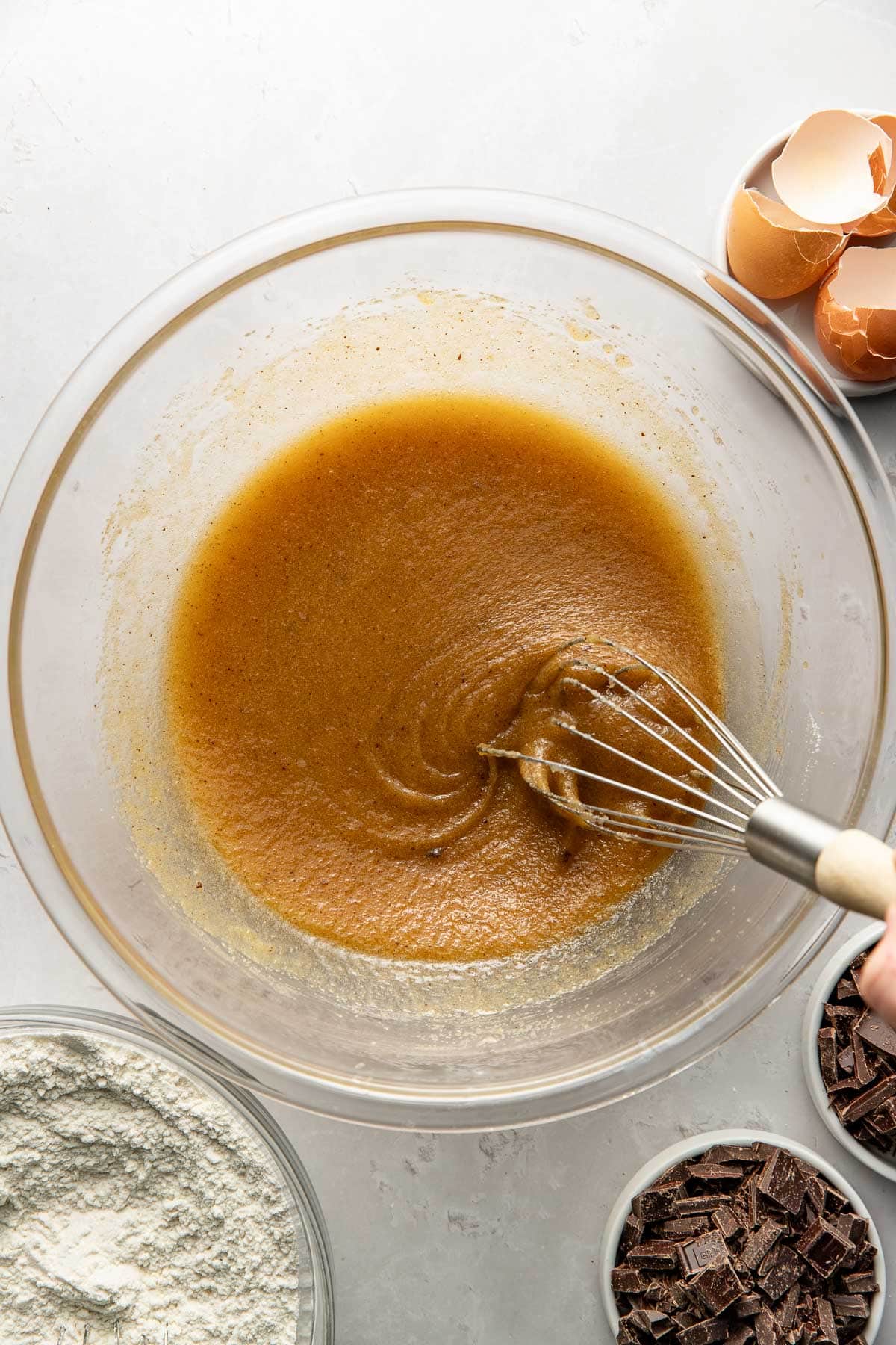 A hand whisking a golden-brown batter in a glass bowl, surrounded by bowls of flour, chopped chocolate, and empty eggshells on a light countertop.