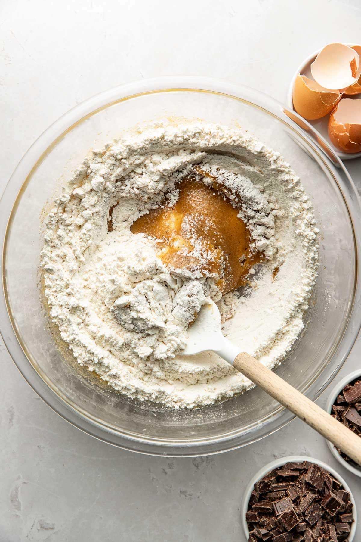 A glass mixing bowl with flour, wet batter, and a spatula. Nearby are cracked eggshells in a bowl and a bowl of chopped chocolate pieces on a light surface.