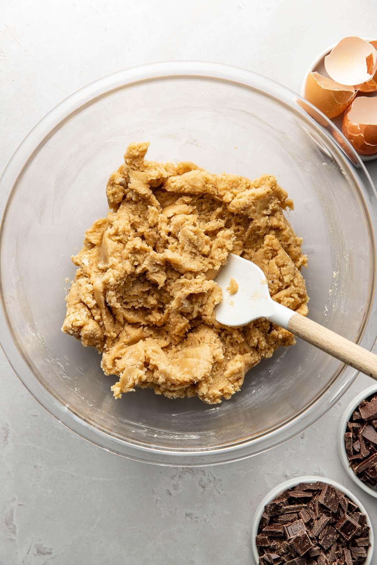 A glass bowl filled with cookie dough and a white spatula, surrounded by small bowls of chocolate chunks and broken eggshells on a light countertop.