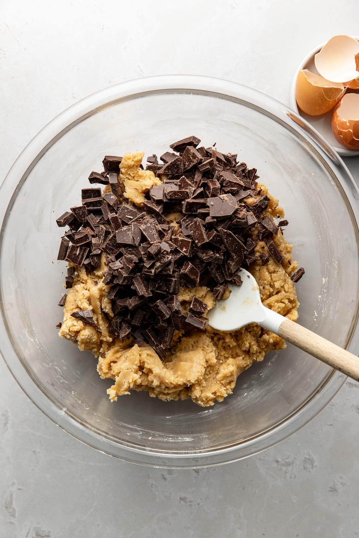 A glass bowl filled with cookie dough and chopped dark chocolate, being mixed with a white spatula. In the background, there is a small plate with empty brown eggshells.