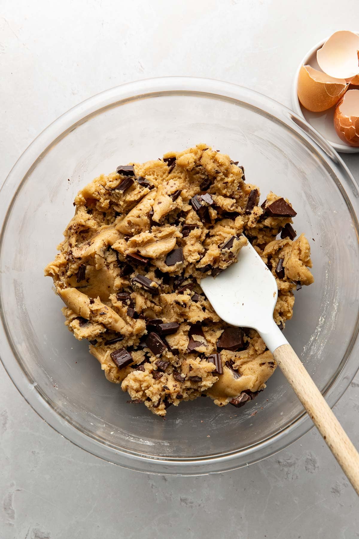 A glass bowl filled with chocolate chip cookie dough and a white spatula. In the corner, there is a small dish with broken eggshells. The background is a light, textured surface.