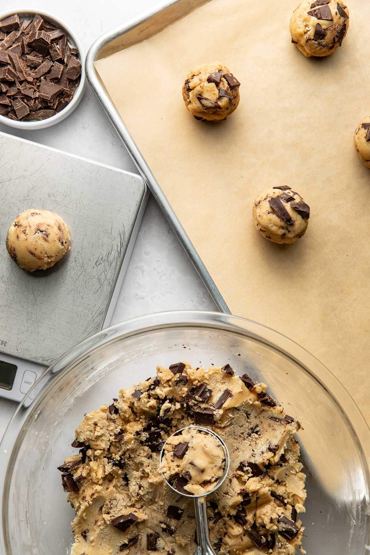 A glass bowl of chocolate chip cookie dough with a scoop rests beside a baking sheet lined with parchment paper, holding several dough balls. Chopped chocolate sits in a small dish nearby.