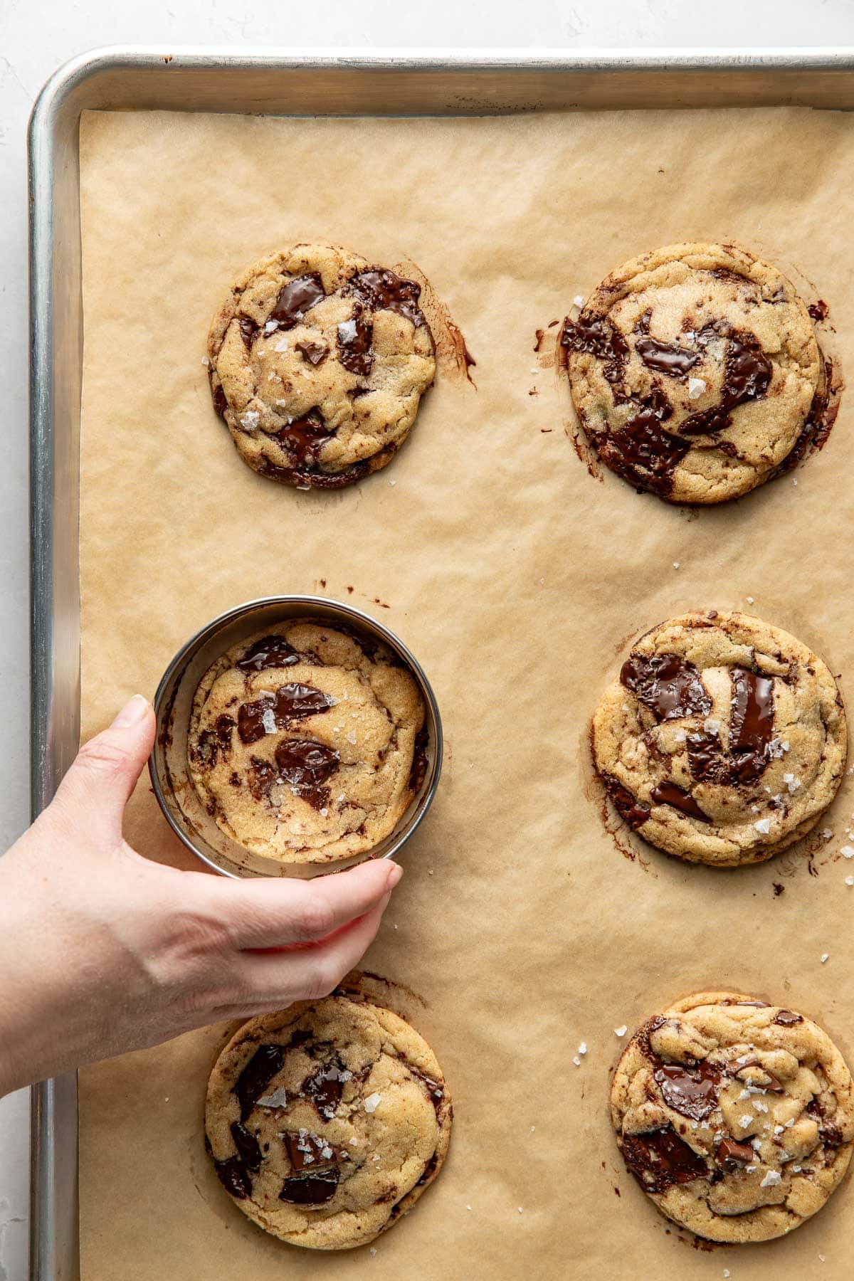 A hand uses a metal ring to shape a freshly baked chocolate chunk cookie on a parchment-lined baking sheet with five other cookies.